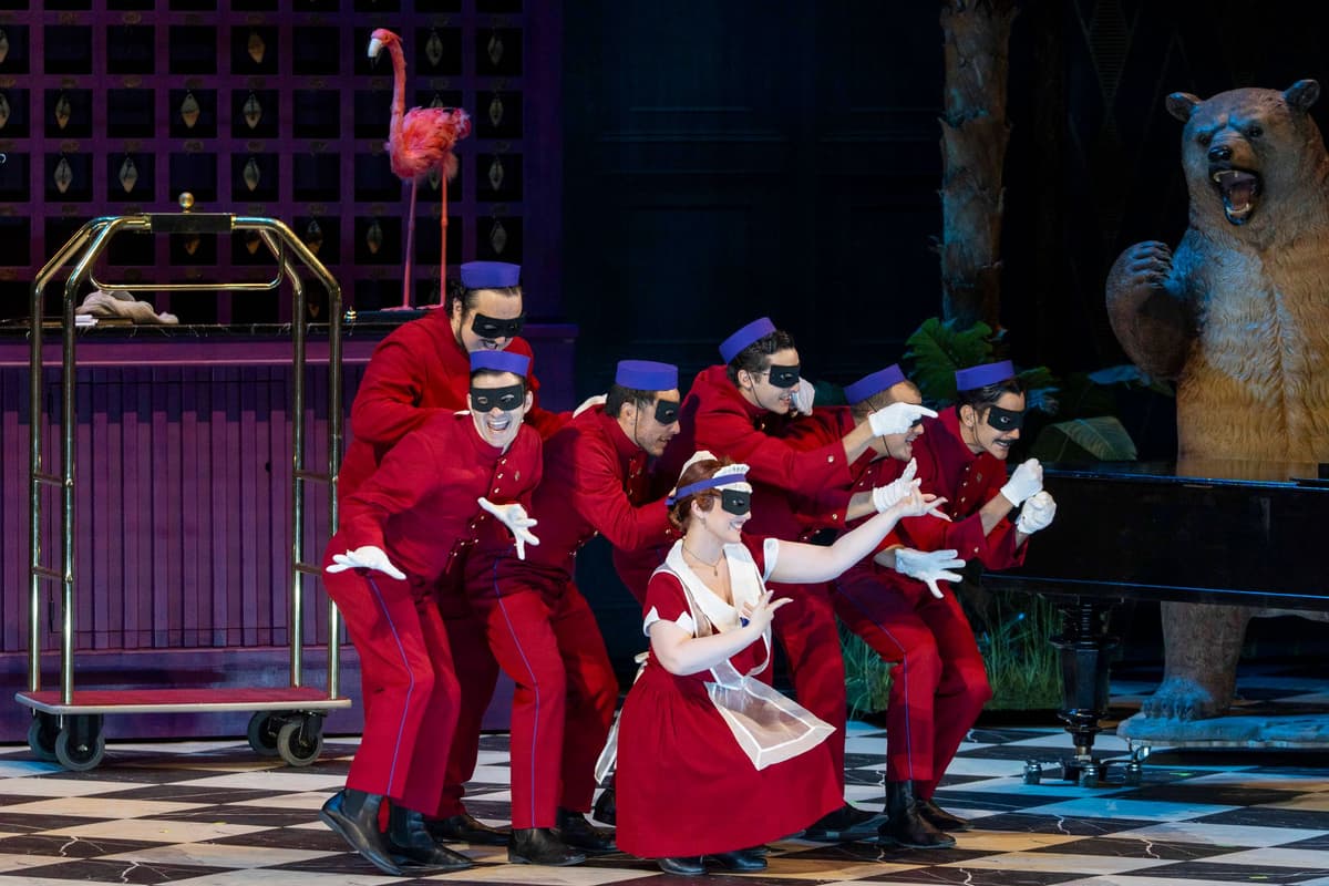 Singers in red bellhop uniforms and eye masks gesture on a hotel-lobby stage set with a flamingo and a bear