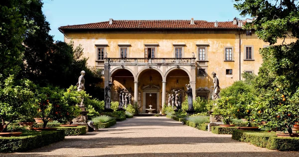 Yellow-ochre palazzo facade with a three-arched loggia and statues, seen across a garden with orange trees